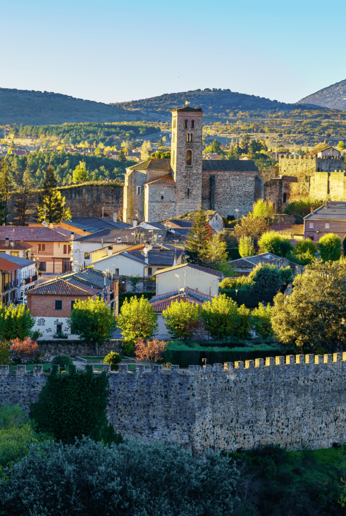 A scenic view of Buitrago de Lozoya village, with medieval city walls in the foreground and green mountains in the background.