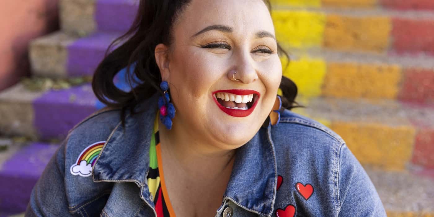 Alysse Dalessandro breaks into a big, natural smile while sitting on a staircase painted with bright rainbow stripes.