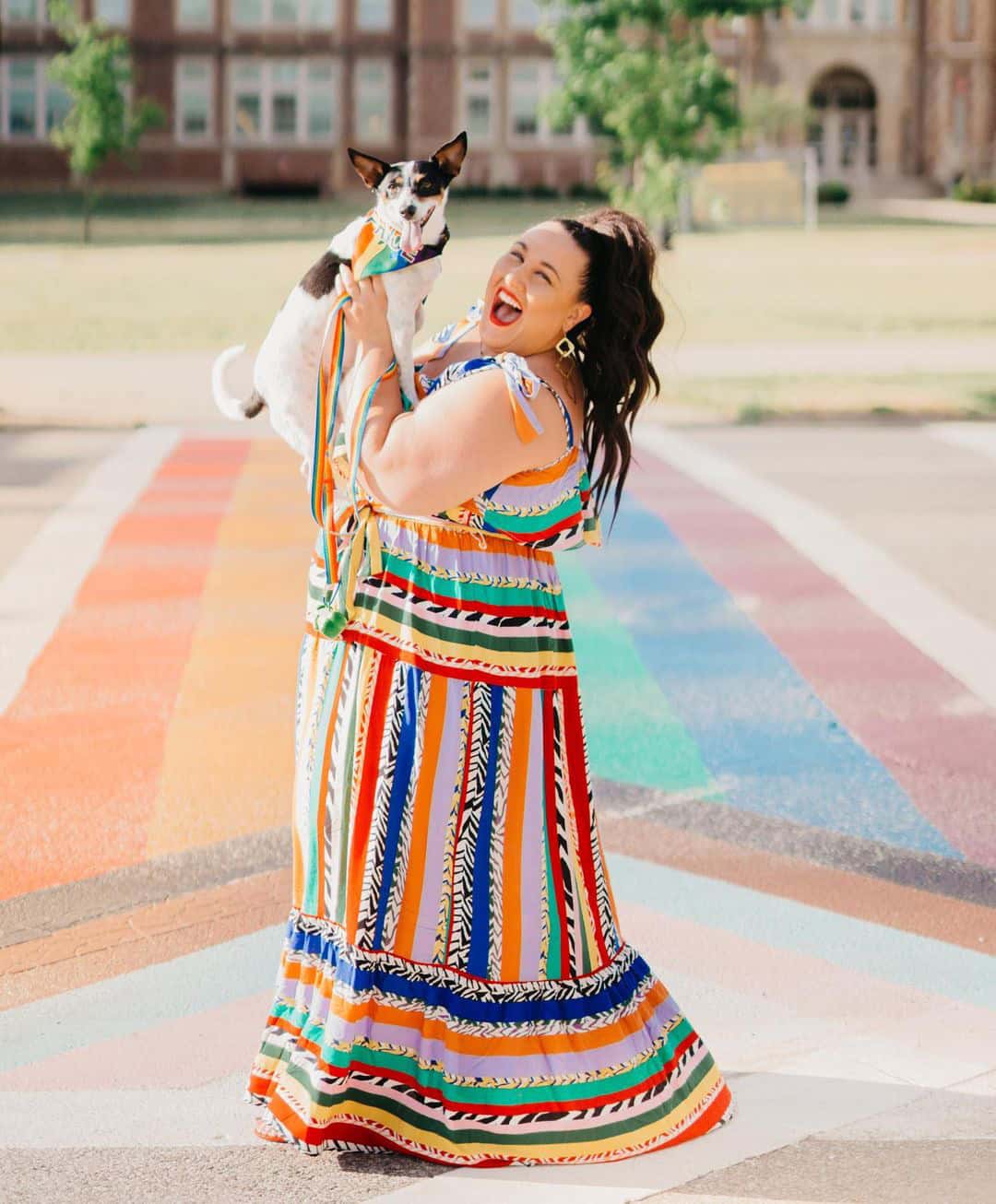 Alysse, in a bright, striped dress, holds up her rescue pup, Bugsy, who is wearing a rainbow-striped neckerchief.