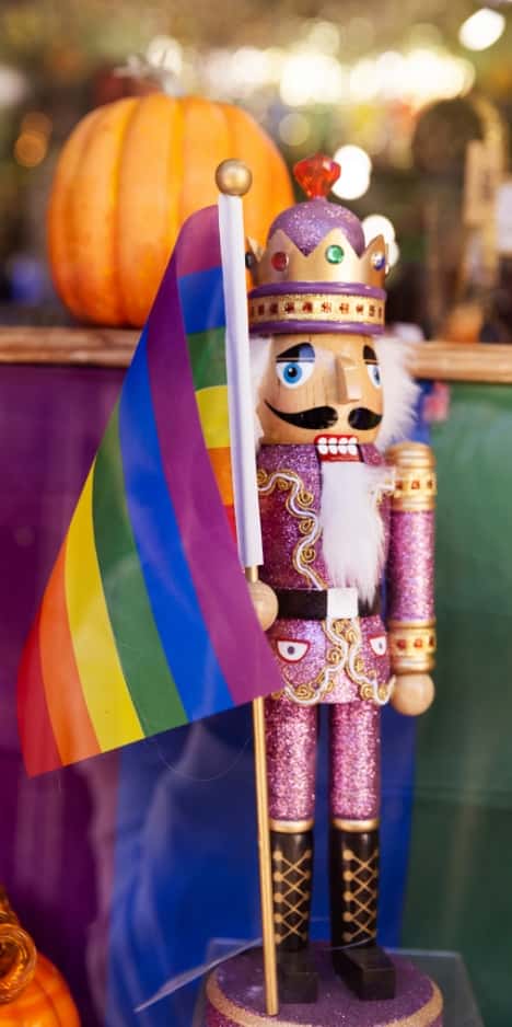 A flamboyant take on a nutcracker soldier, dressed in glittery pink attire and holding a rainbow flag, stands in a shop window.