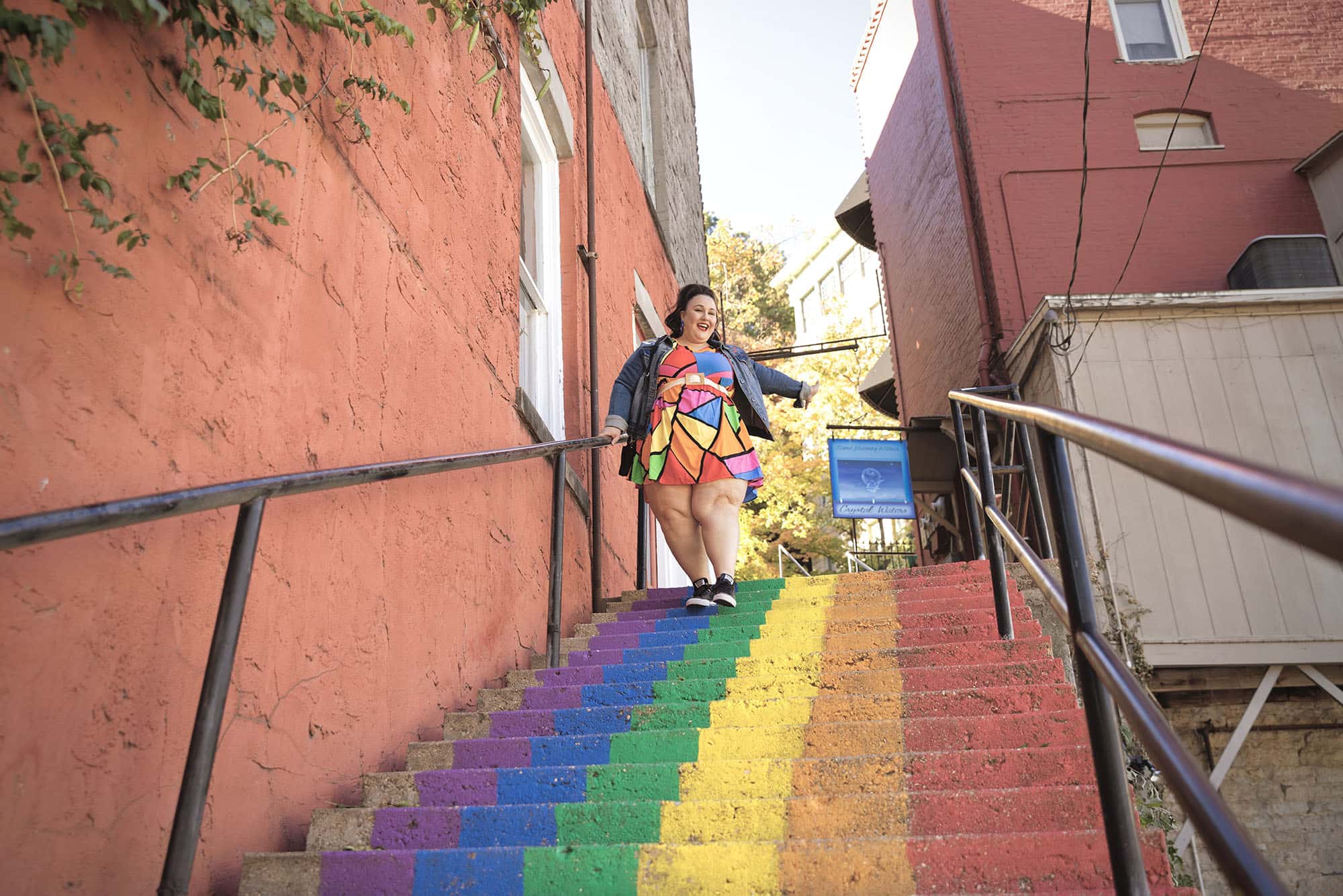 Alysse, in a bright, geometric-patterned, rainbow-colored dress, descends a rainbow-striped staircase.