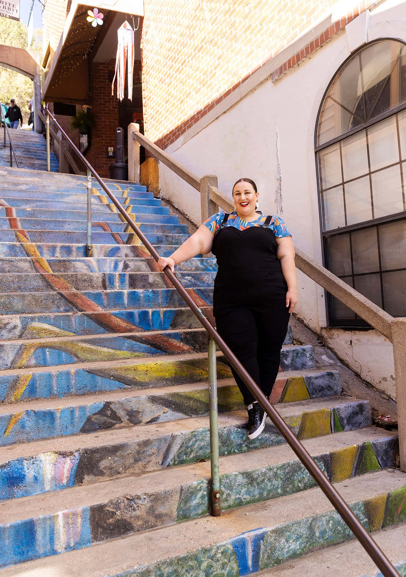 Alysse, in black overalls and a brightly patterned shirt, poses on one of the town’s many painted staircases.