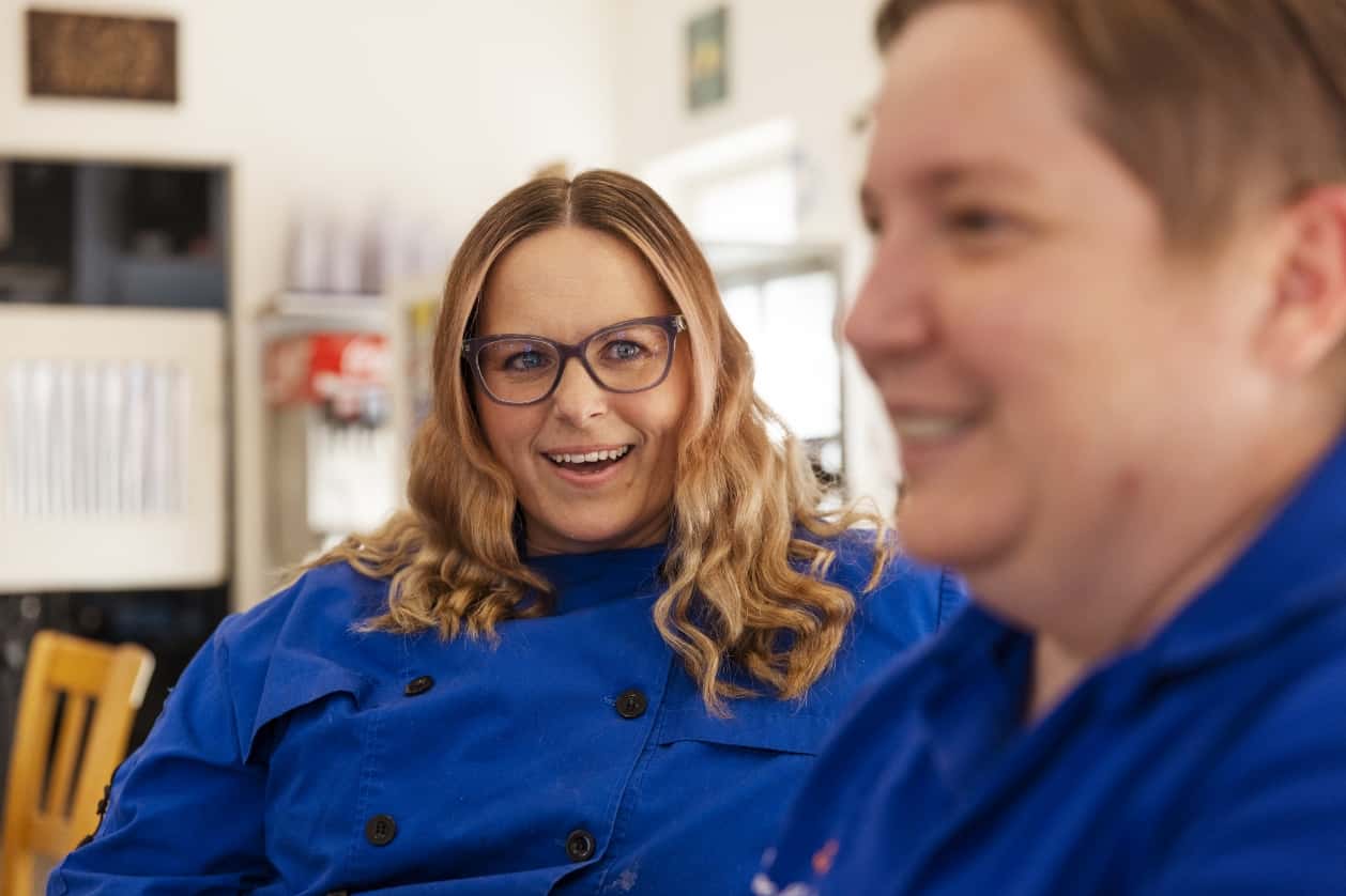 Jennifer McDermott, in a blue chef coat, smiles broadly as Rayna talks with Alysse.
