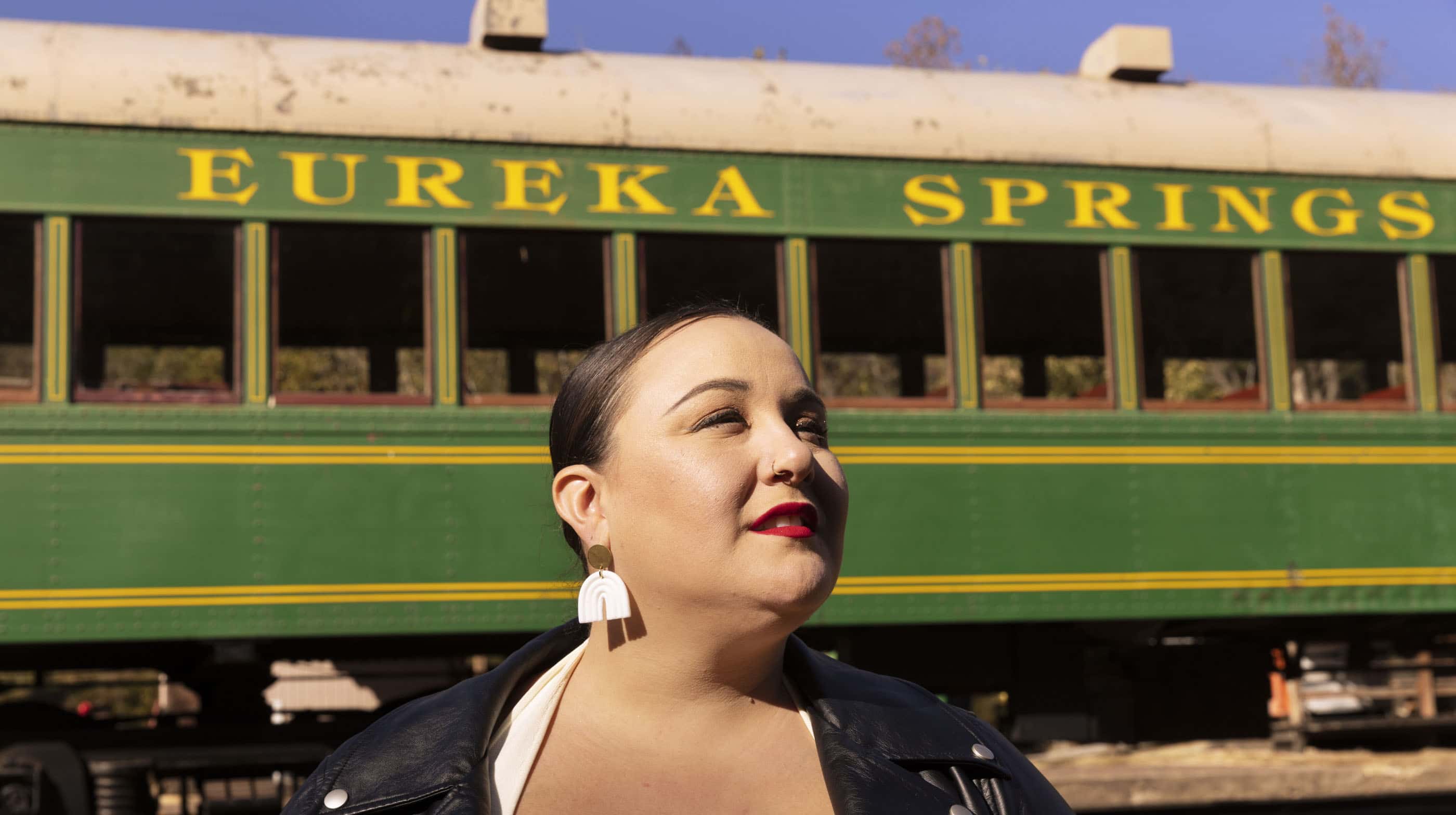 Alysse, wearing white rainbow-shaped earrings, stands in front of a vintage train carriage with its destination, Eureka Springs, painted across it.