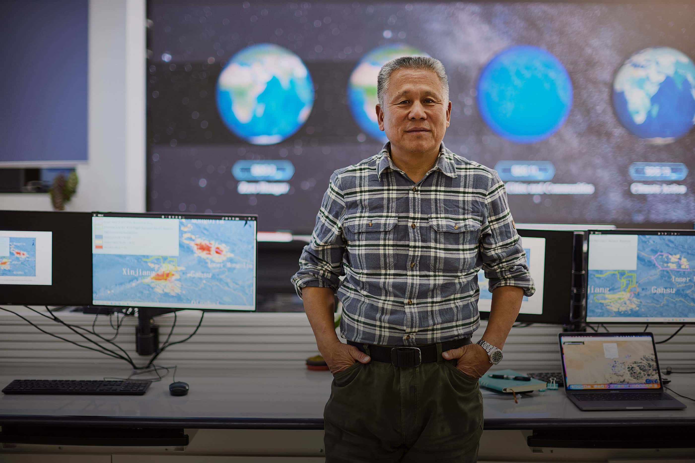 Liu leans against a table in his imaging lab. Computer monitors show satellite images of Earth.