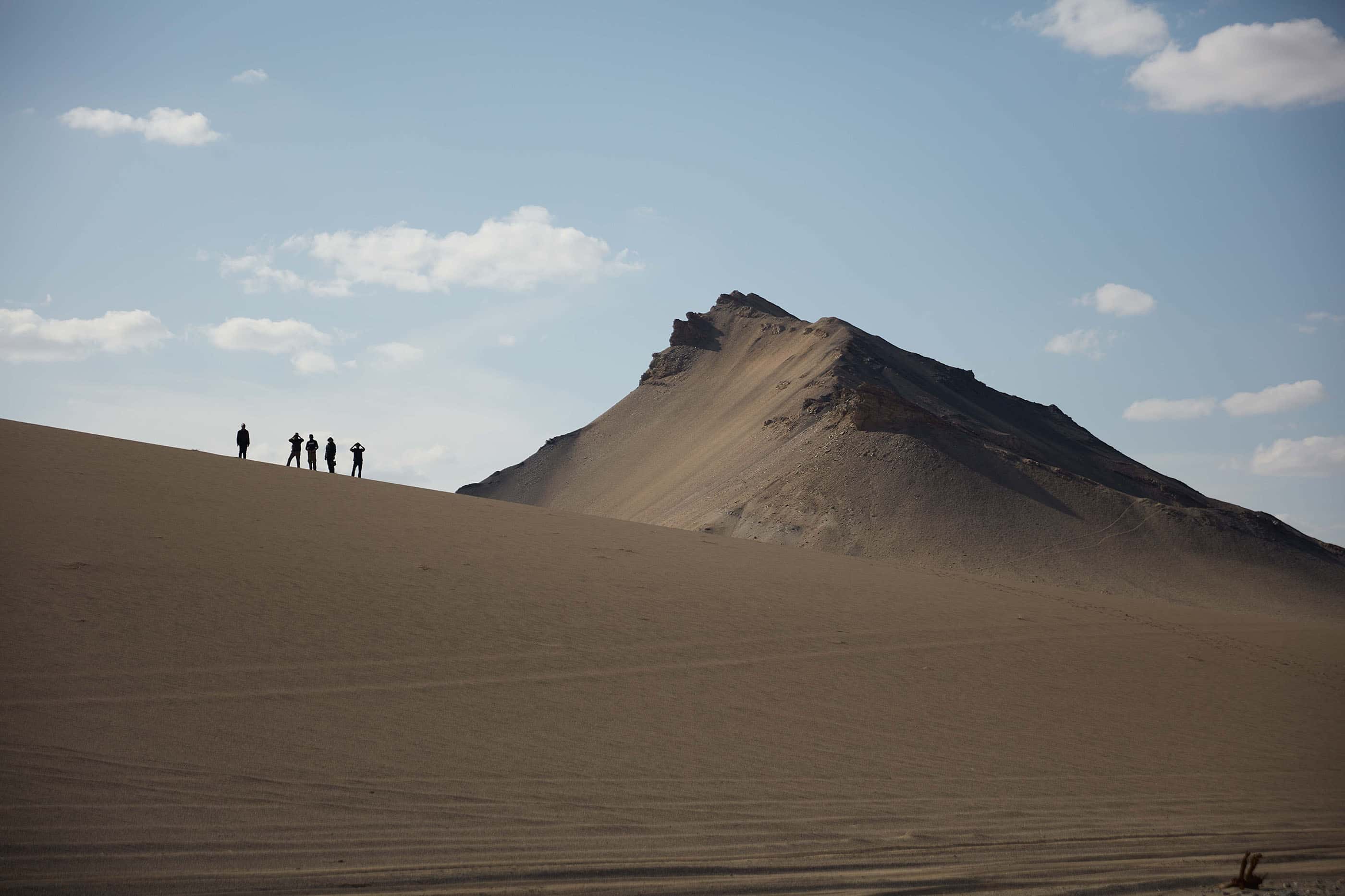 A towering sand dune in the Gobi Desert, with five people visible in the distance.