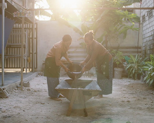 Two individuals working together with a bowl at an outdoor table, surrounded by plants and bathed in soft sunlight.