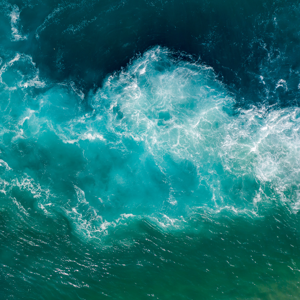 Aerial view of ocean waves with varying shades of blue and green, creating textured patterns from the frothy white water.