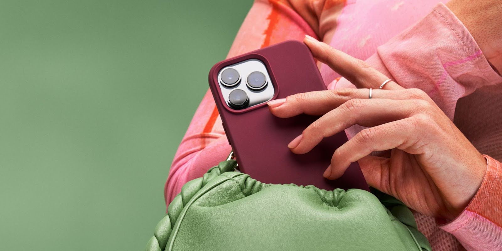 A person in a pink and orange top holds a smartphone with a maroon case above a green, cinched fabric bag against a green background.