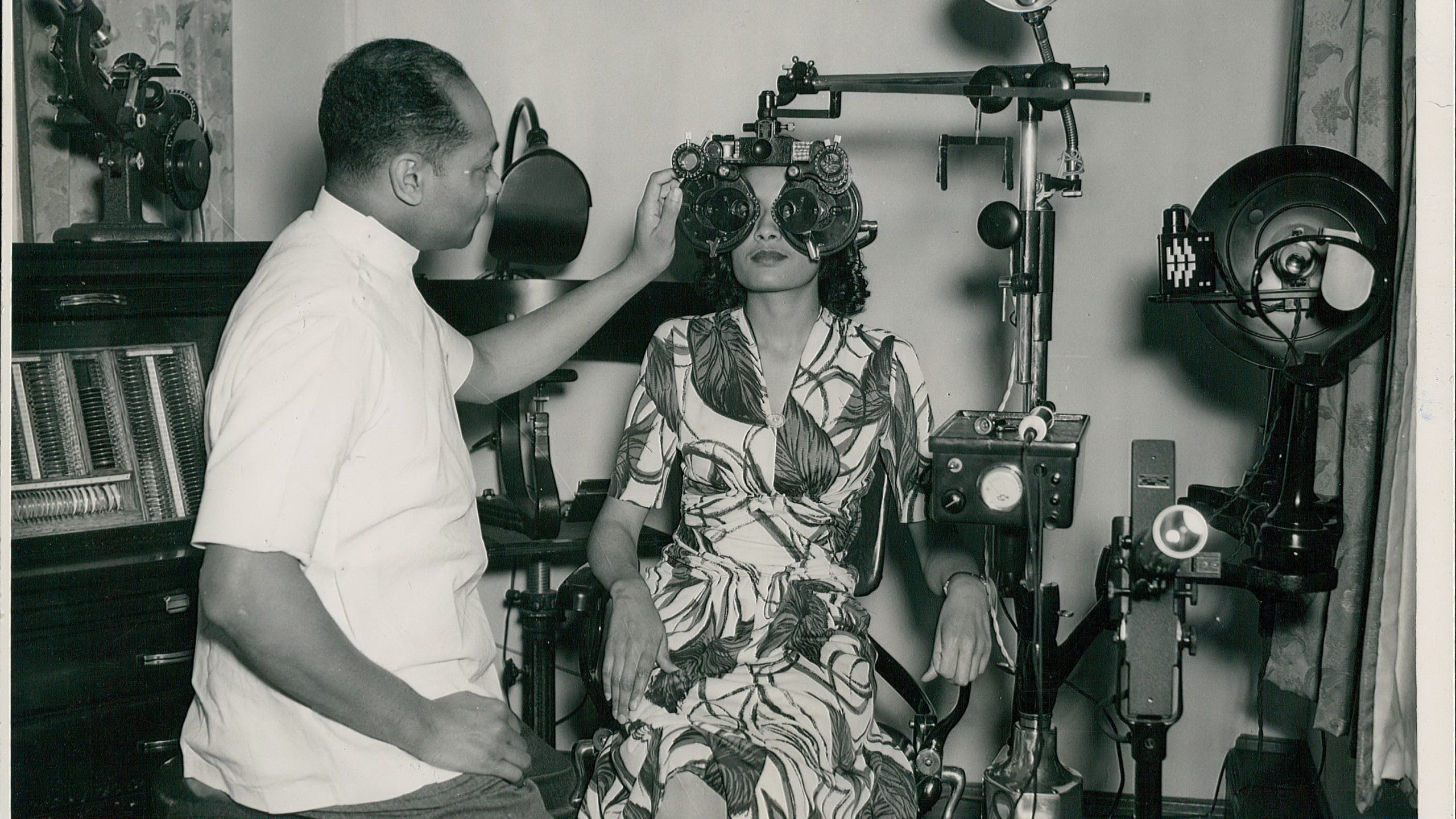 A person in a floral dress undergoing an eye exam, seated at a phoropter, while a technician adjusts the machine.