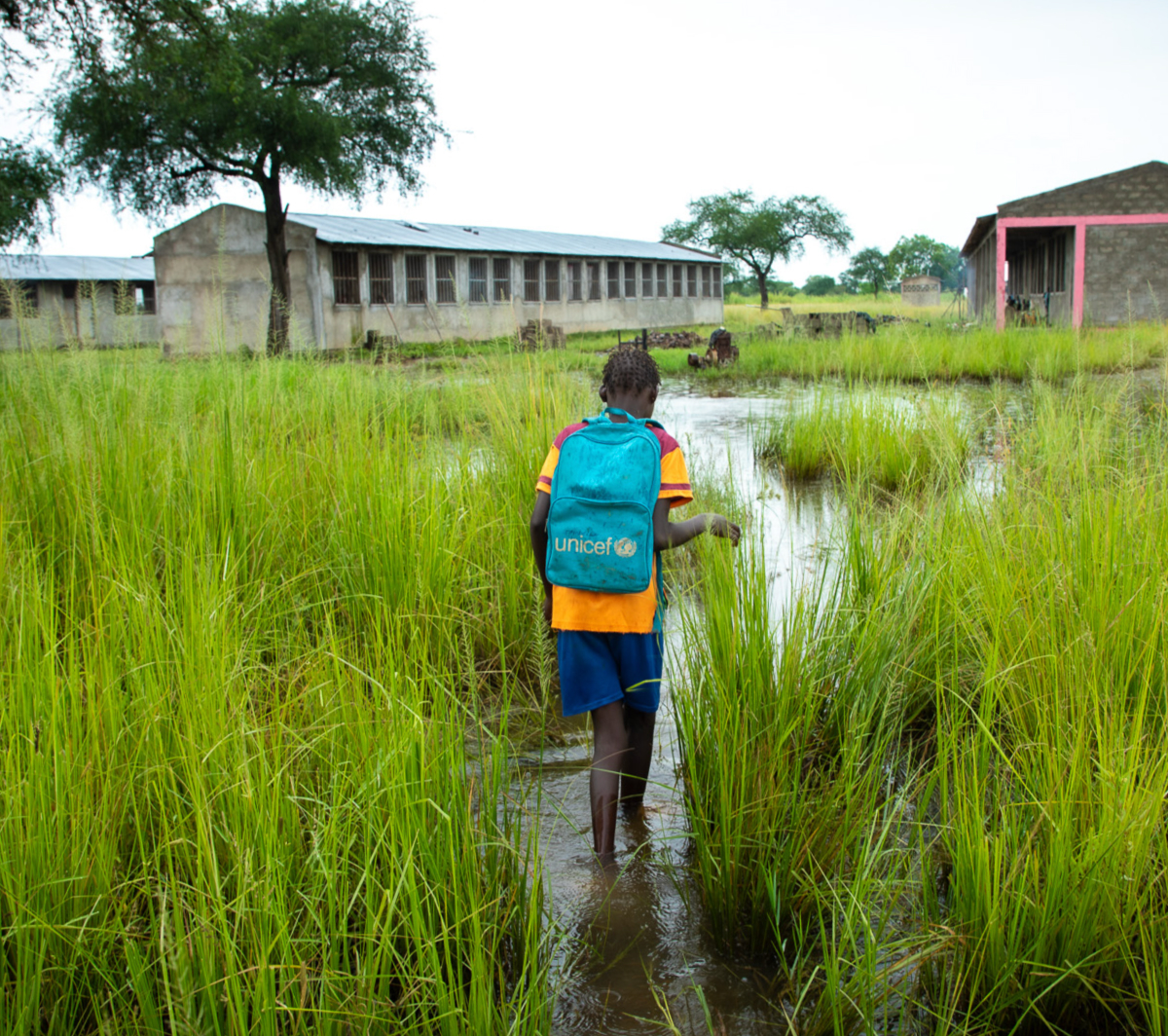 A child with a UNICEF backpack walks through a flooded grassy area towards buildings in the distance.