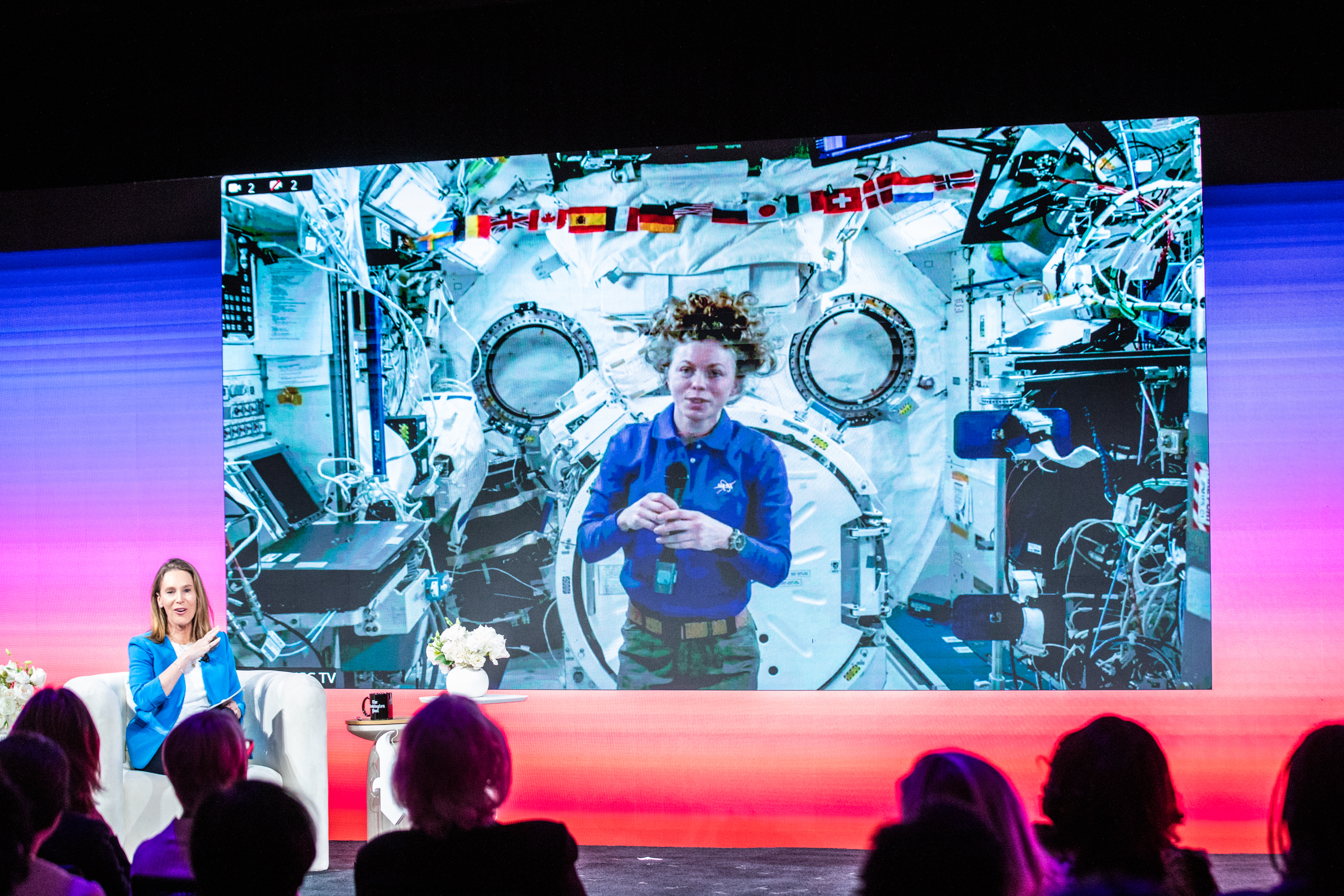 A woman on stage speaks to an audience while a large screen behind her shows an astronaut inside a space station module.
