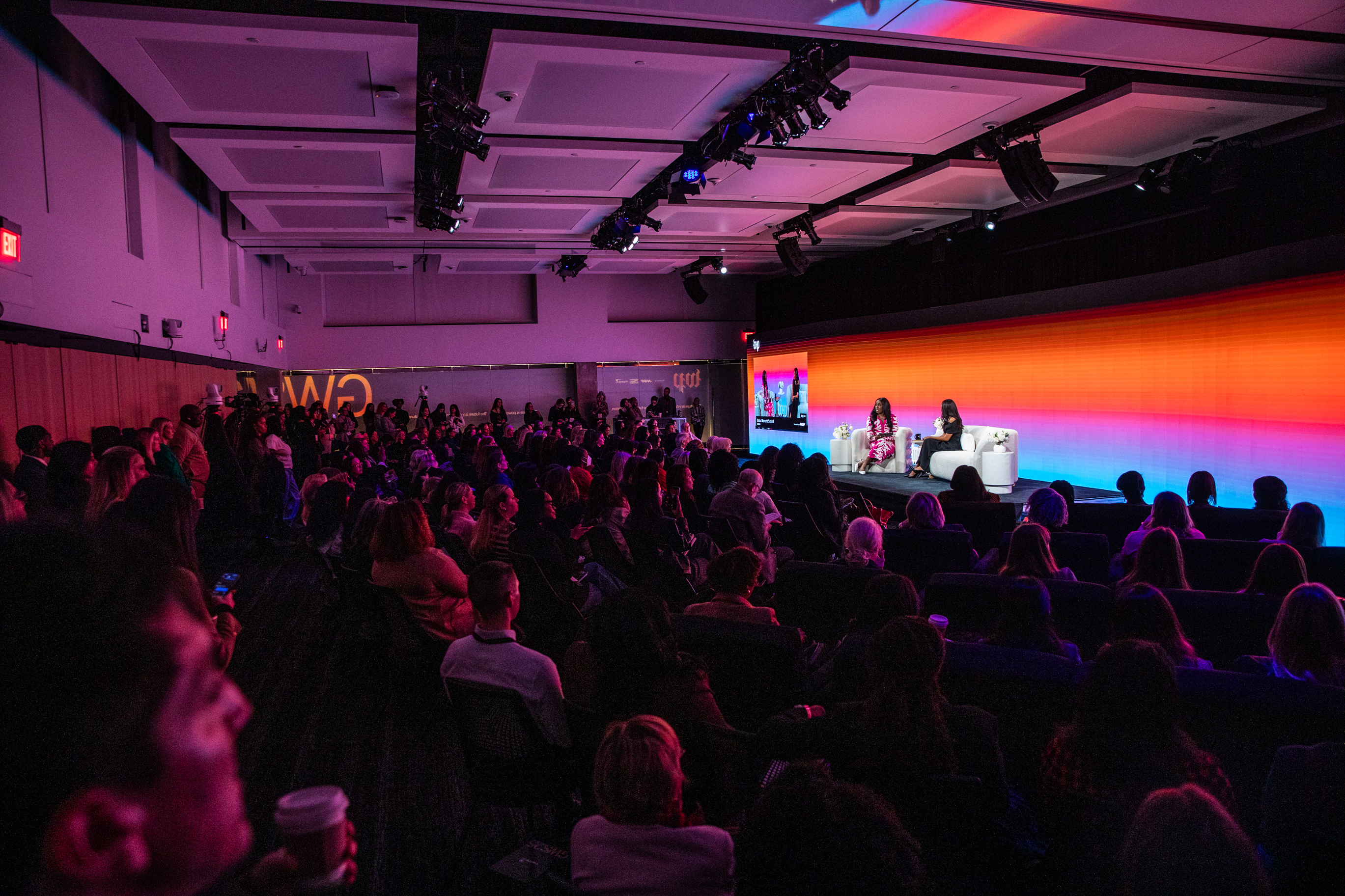 A large audience sits in a dimly lit auditorium watching two speakers in conversation on stage, with a colorful gradient screen behind them.