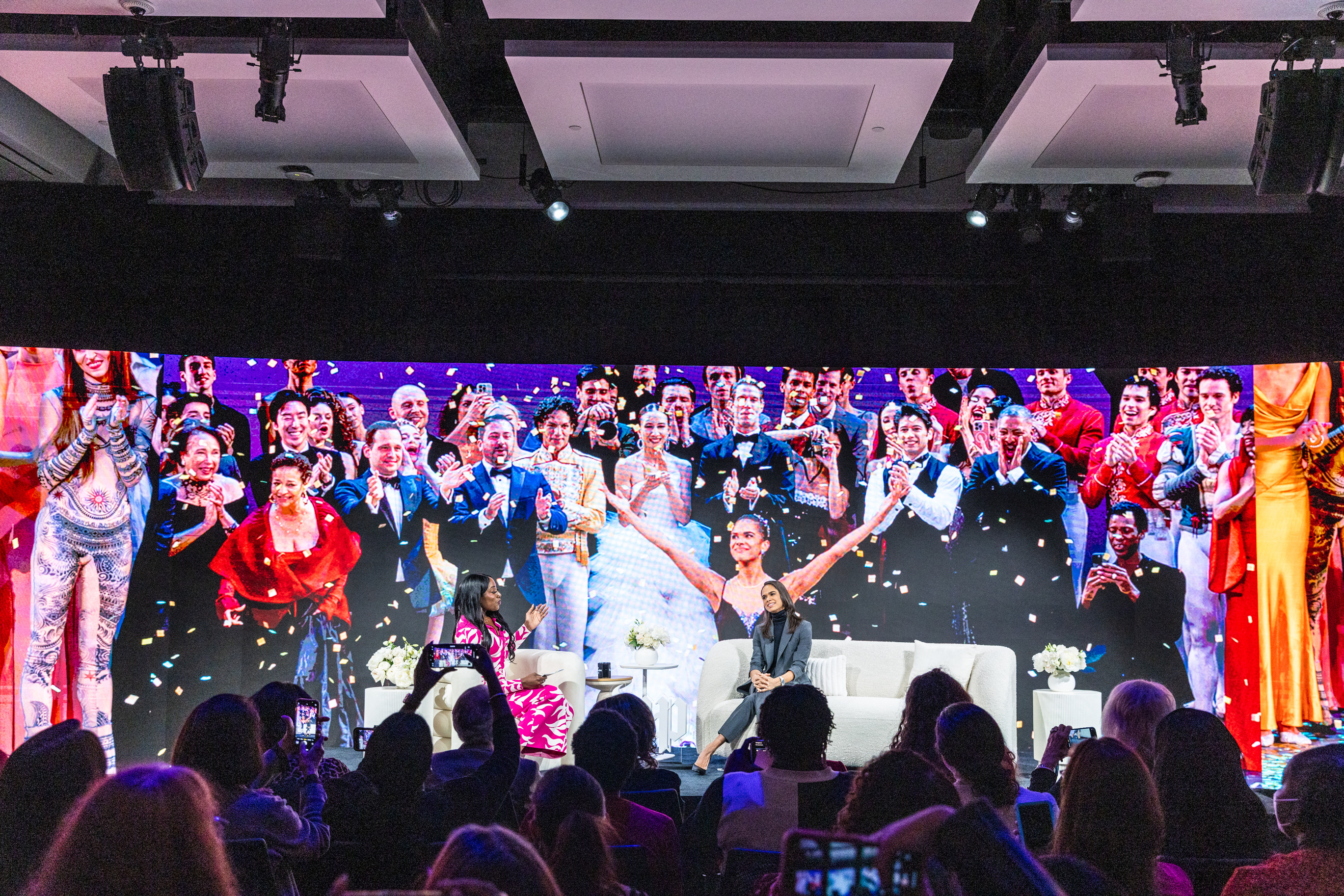 A person sits on stage for a talk in front of an audience, with a large screen behind them displaying a festive group celebrating.