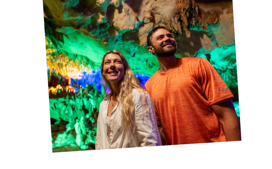 Two people stand in an illuminated cave with colorful green and orange lights highlighting rock formations in the background.