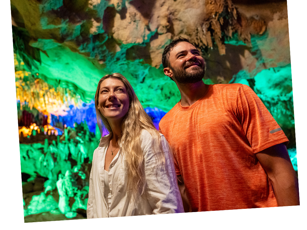 A woman and a man stand inside a cave with colorful lighting, looking up and smiling; stalactites and rock formations are visible in the background.