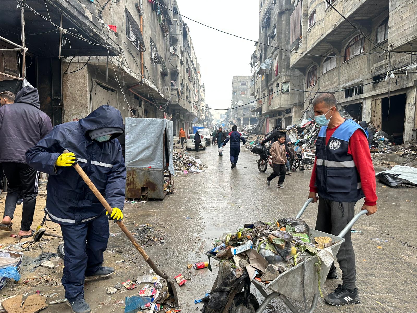 Workers in protective clothing clean debris and trash from a littered street in a damaged urban area, while people walk and ride bicycles in the background.
