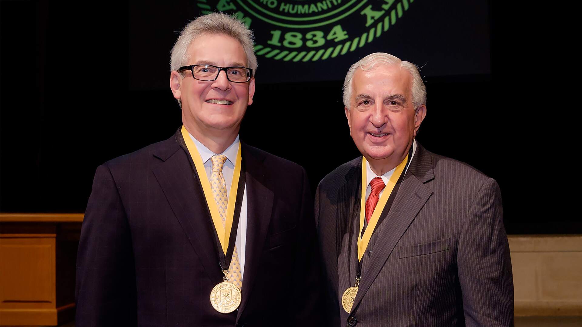 Two men in suits, each wearing a gold medallion on a black ribbon around their necks, stand in front of a large emblem projected on a screen.