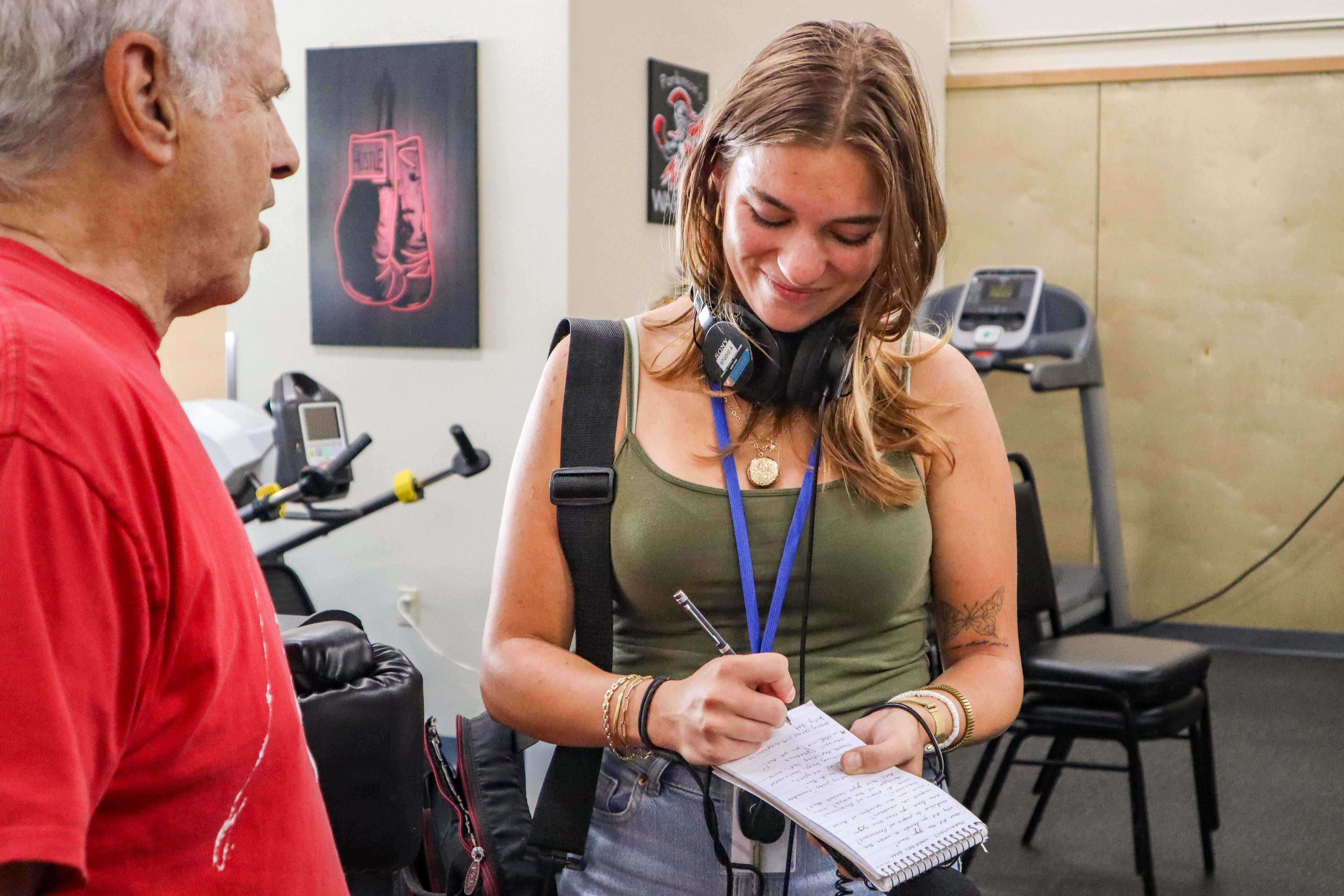 A young woman with headphones around her neck writes in a notebook while an older man speaks to her in a gym with exercise equipment in the background.