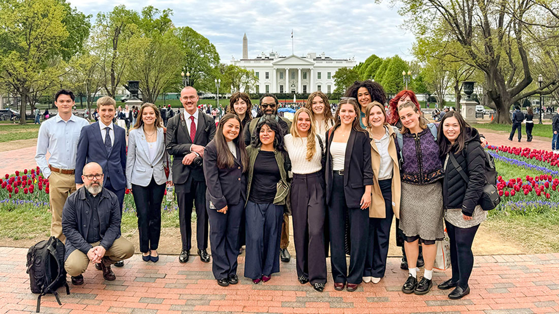 A group of people pose for a photo in front of the White House, with the Washington Monument visible in the background and tulips in bloom nearby.