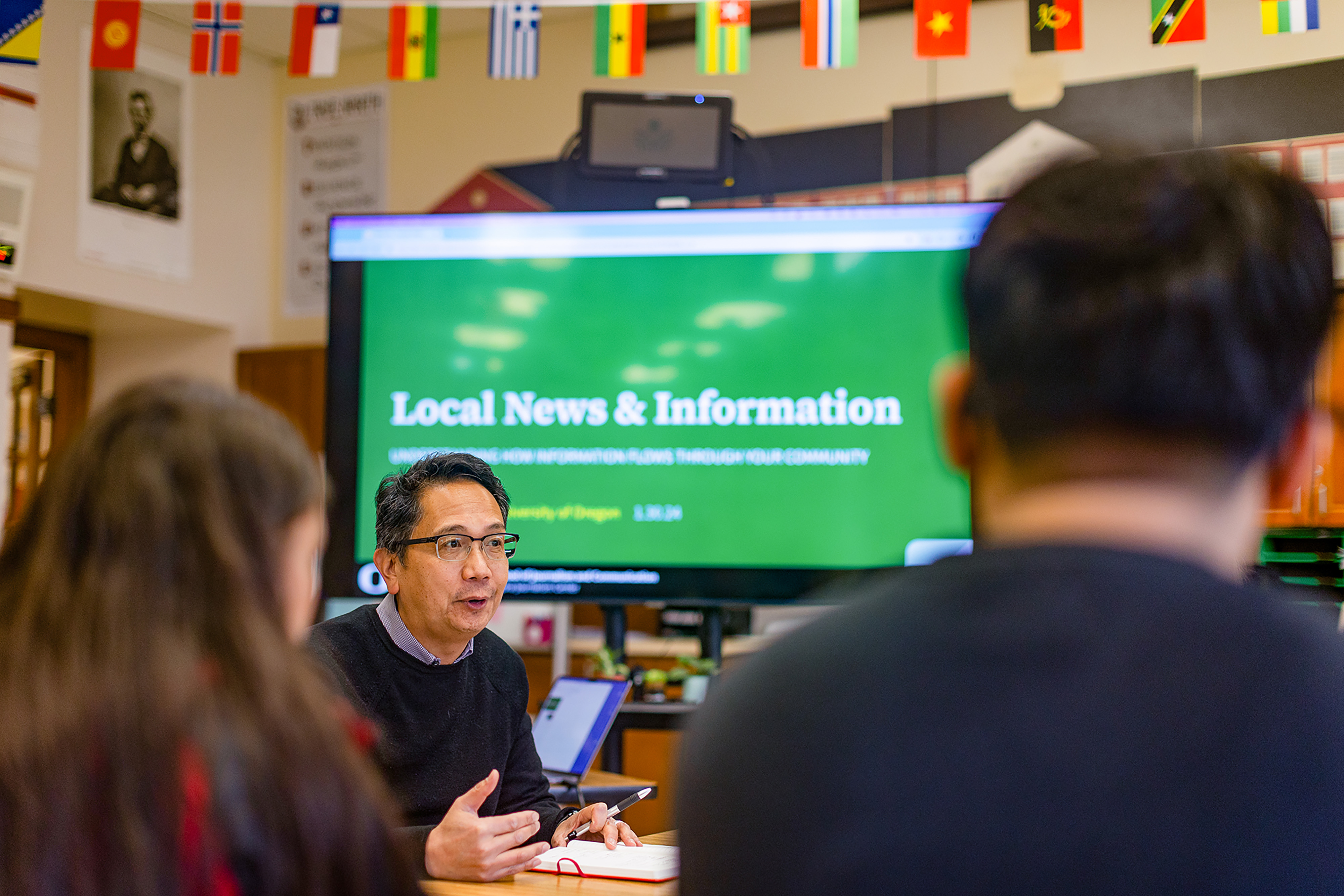 A man speaks to two people in a classroom with international flags and a large screen displaying "Local News & Information" in the background.