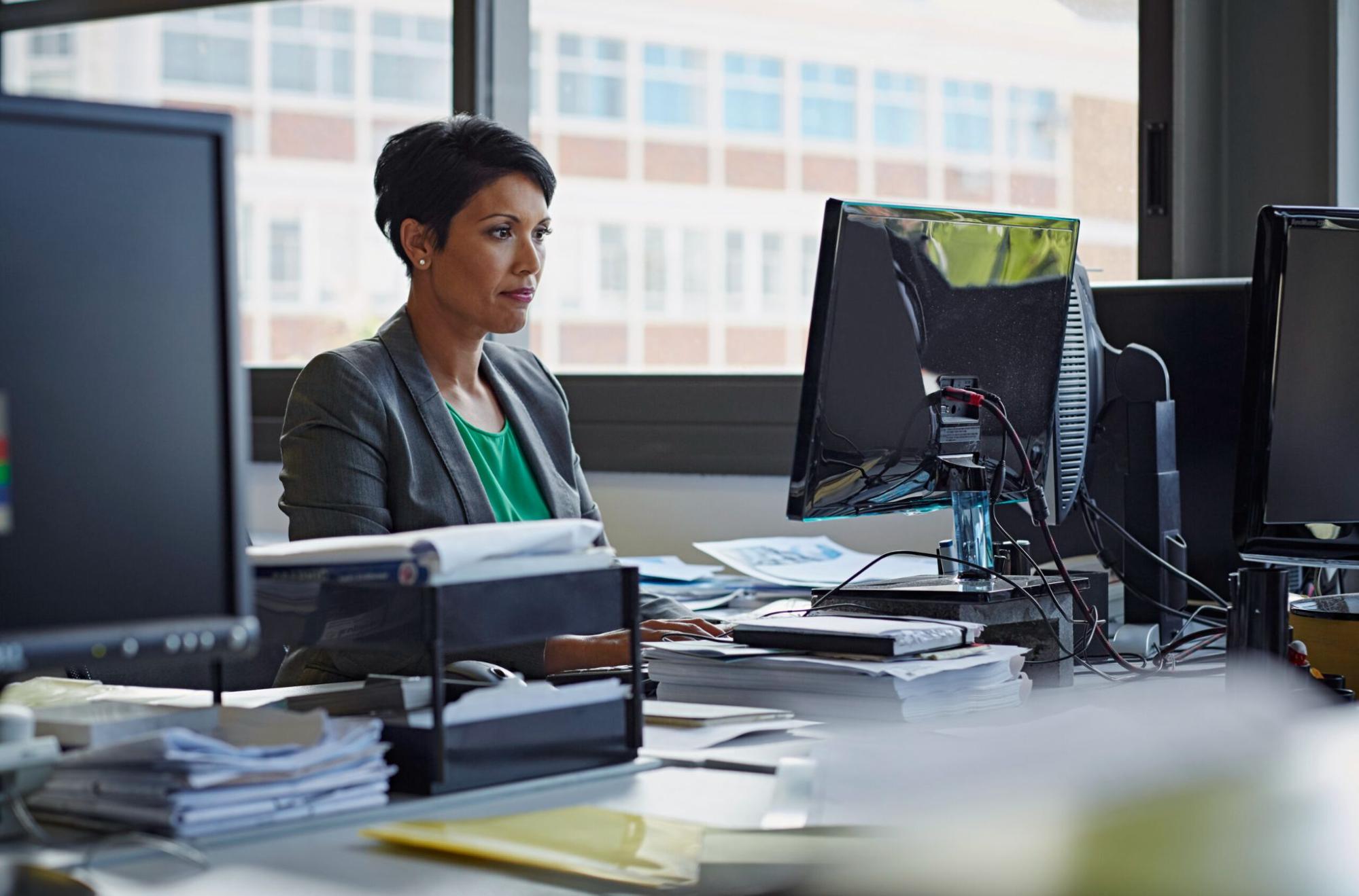 A woman in business attire sits at a desk working on a computer in an office with paperwork and office supplies around her.