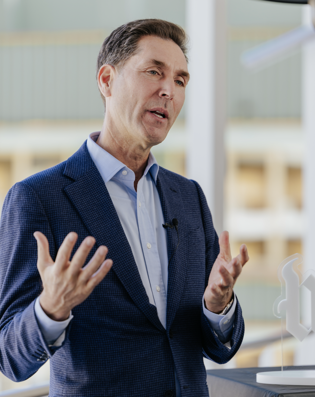 A man in a blue suit and light blue shirt speaks with hand gestures. He wears a clip-on microphone and stands indoors with blurred windows in the background.