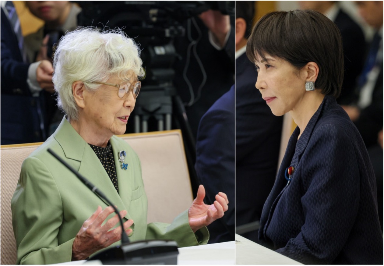 Two women sit across from each other at a table, engaged in conversation during a formal meeting, with microphones and cameras visible in the background.