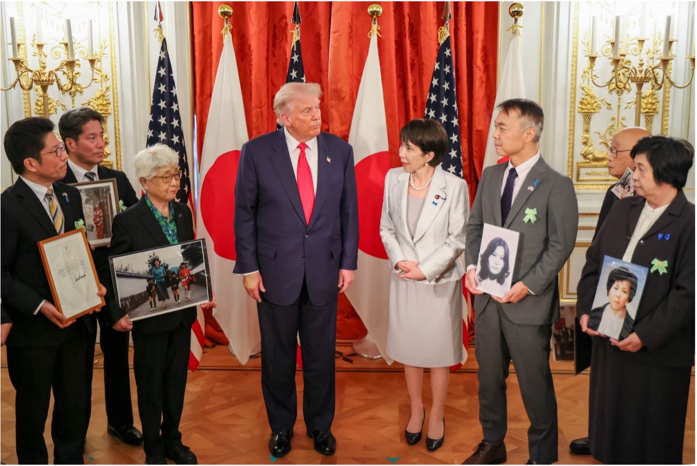 Donald Trump stands with a group of people holding framed photos, with US and Japanese flags and red curtains in the background.