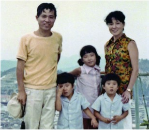 A family of five poses outdoors; two adults stand behind three young children, with a landscape visible in the background.