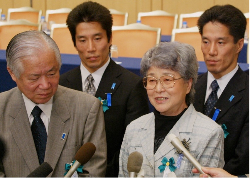 A group of four people in business attire, three men and one woman, stand together with blue ribbons, speaking into microphones at a press event.