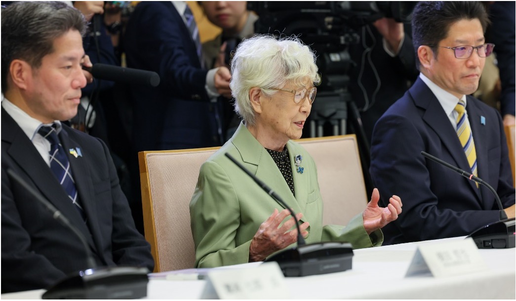 An elderly woman speaks seated between two men in suits at a conference table, with microphones and cameras in the background.