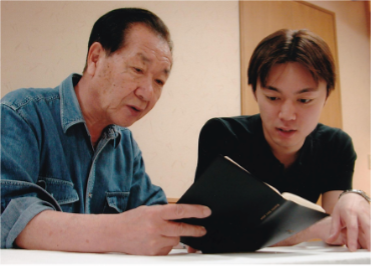 Two men sit at a table, closely reading and discussing the contents of an open book together.