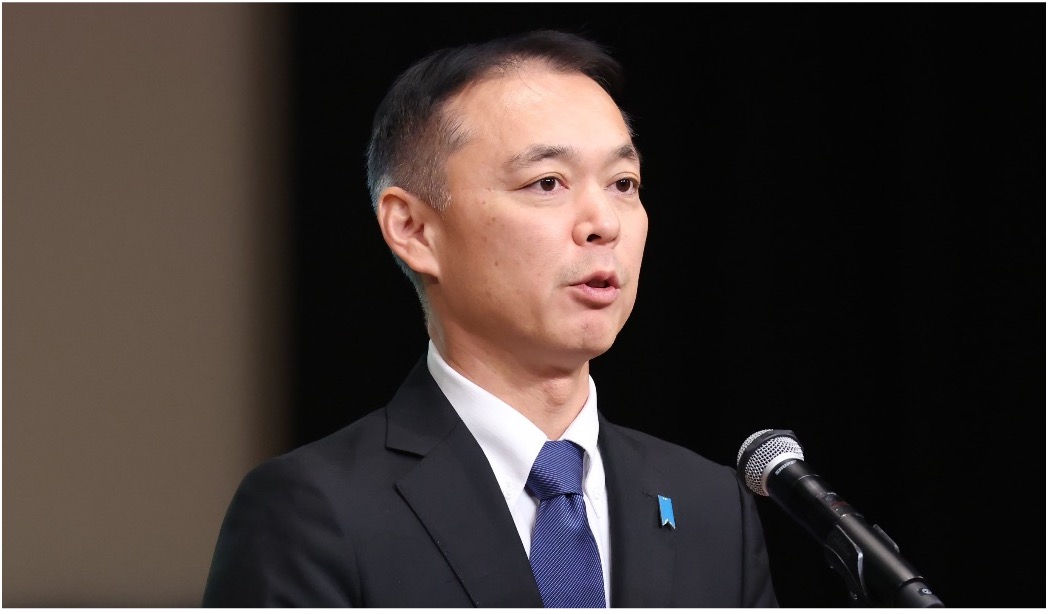 A man in a suit and tie speaks into a microphone at an event, standing in front of a dark background.