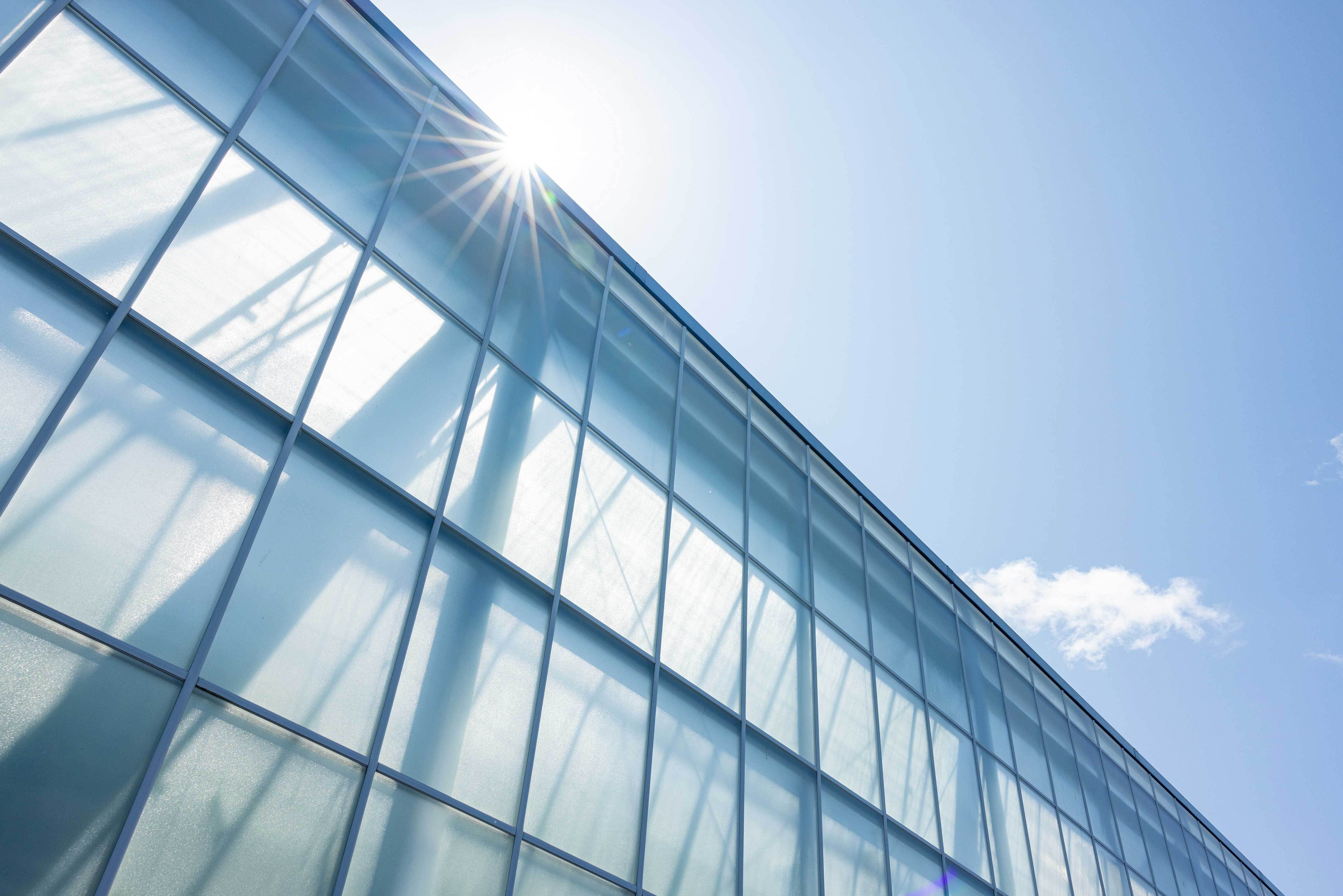 Sunlight shines through the glass panels of a modern building facade, with a clear blue sky and a small cloud in the background.