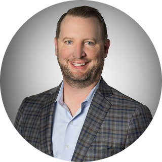 A man with short light brown hair and a trimmed beard, wearing a blue dress shirt and a plaid blazer, smiles at the camera against a gray background.