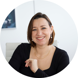 A woman with shoulder-length brown hair, wearing a black top and pearl earrings, is smiling while seated in an office setting.