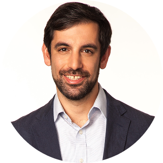 Man with short dark hair and beard wearing a navy jacket and light-colored shirt, smiling at the camera against a plain background.