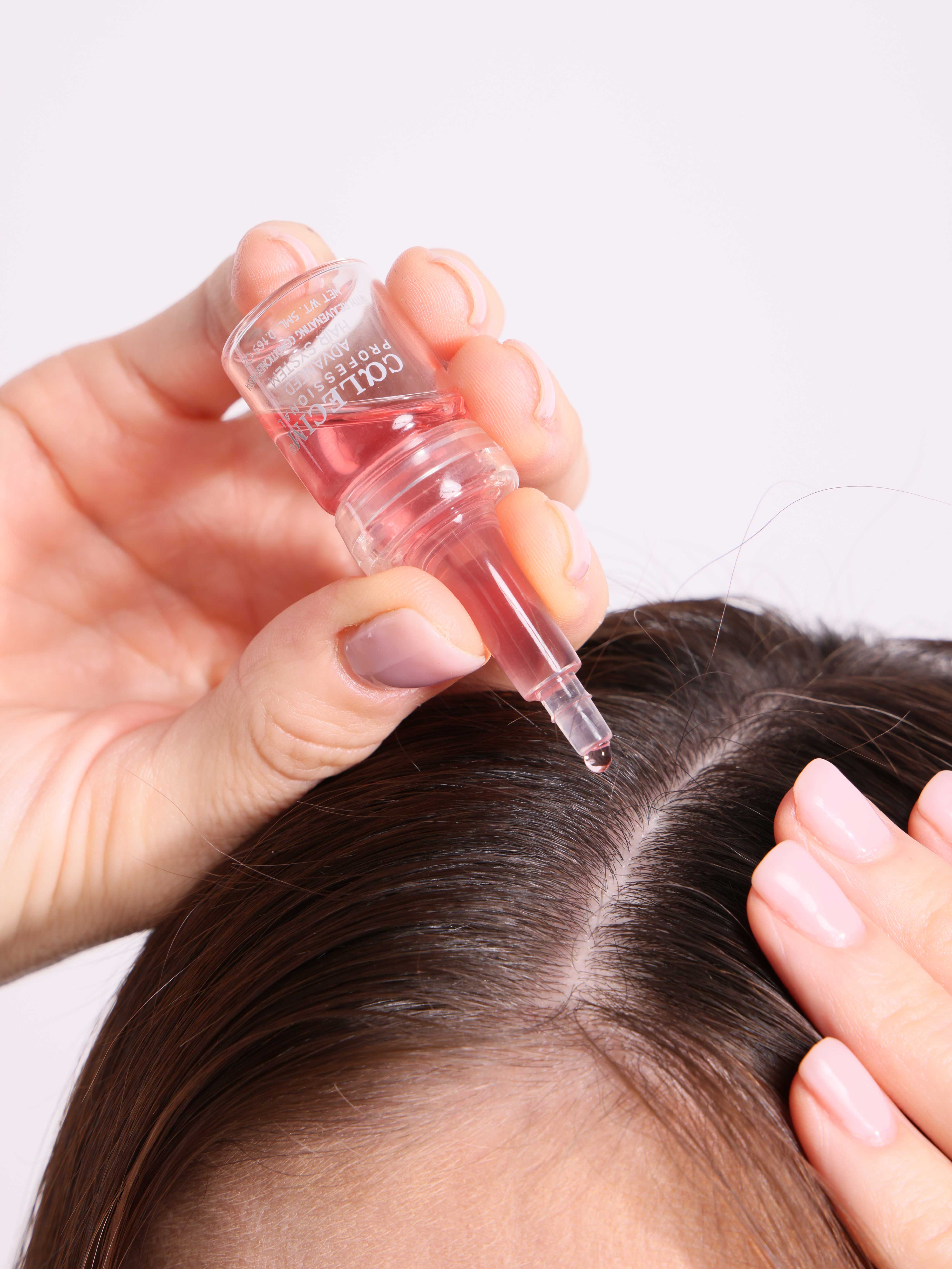 A hand applies a dropper of pink liquid to the scalp of a person with brown hair.