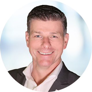 Smiling man with short dark hair wearing a white collared shirt and dark blazer against a light blurred background.
