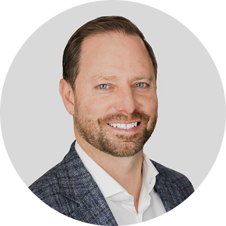 A man with short brown hair and a beard, wearing a white shirt and patterned blazer, smiling at the camera against a light gray background.