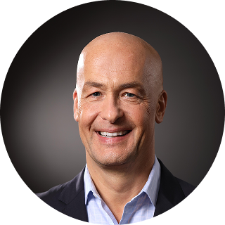 A smiling middle-aged man with a bald head, wearing a dark blazer and light blue shirt, is posed against a dark gradient background.