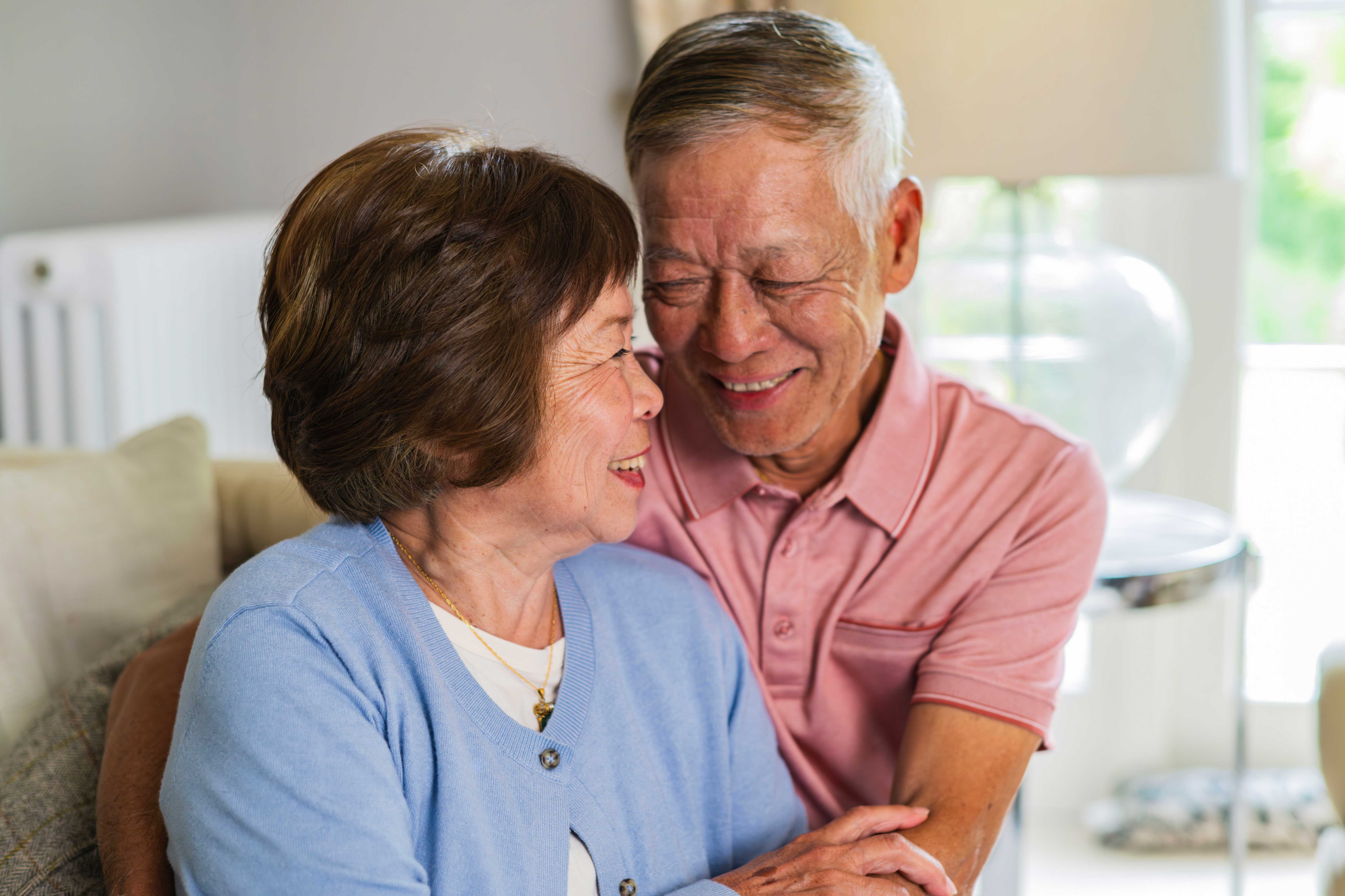An elderly couple sits close together on a couch, smiling and embracing warmly in a bright, comfortable living room.