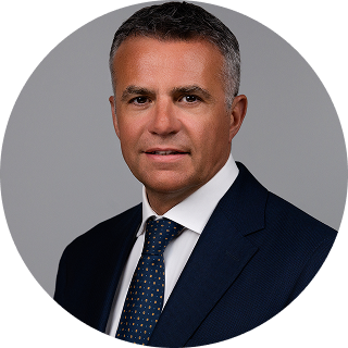 Man in a dark suit and dotted tie poses for a formal headshot against a plain gray background.