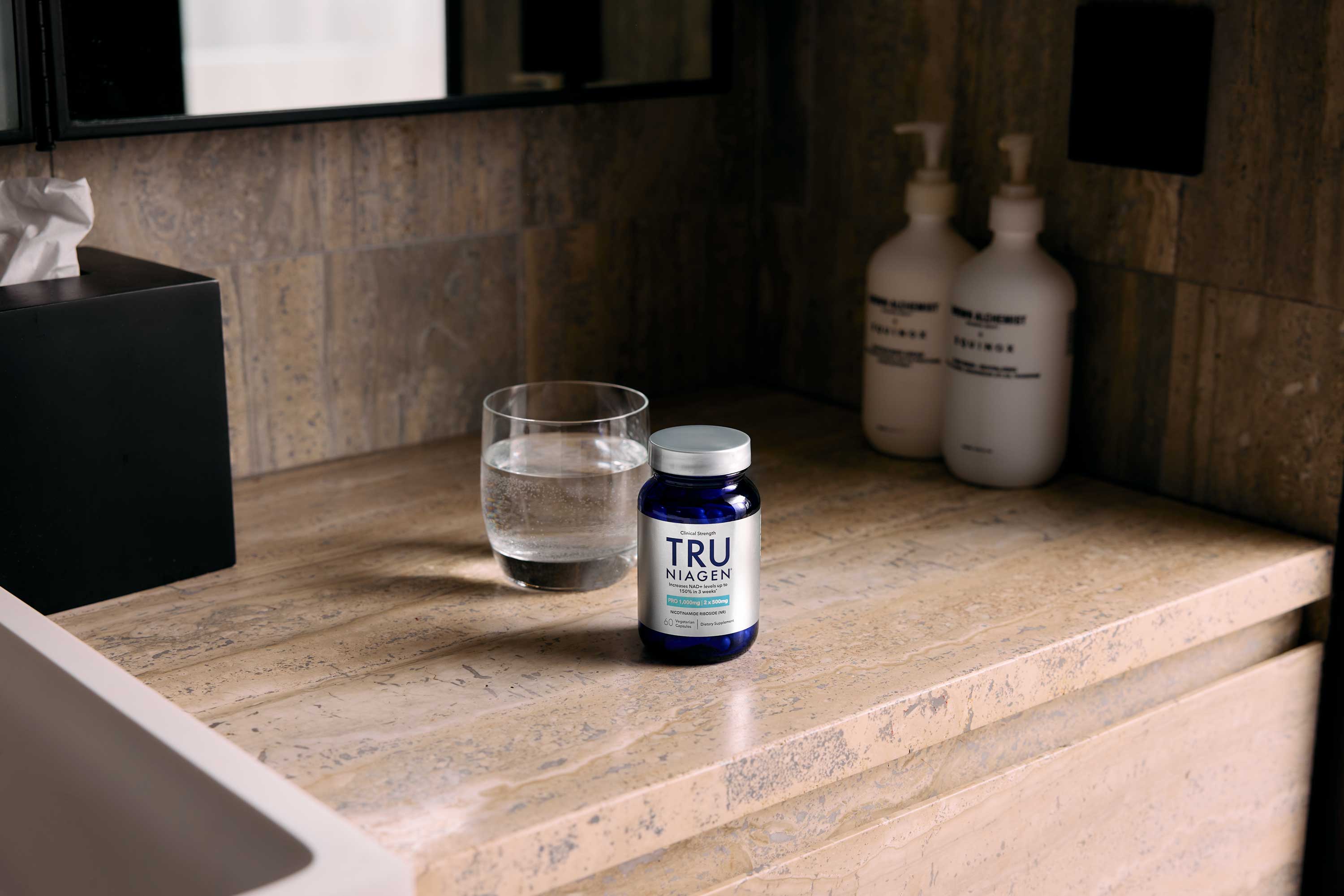 A bottle of Tru Niagen supplements, a glass of water, and two pump bottles are placed on a stone bathroom countertop next to a sink.