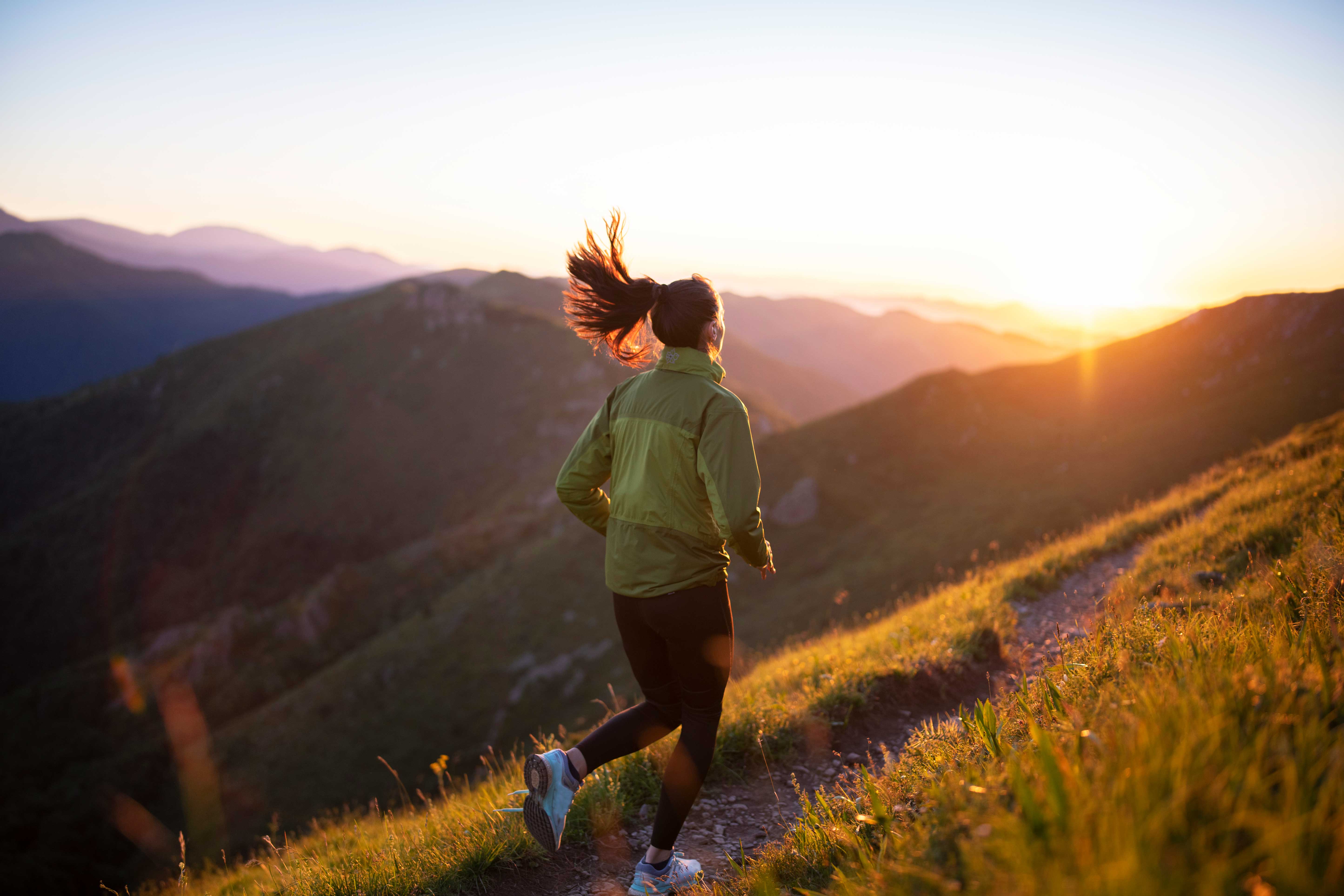 A person runs along a grassy mountain trail at sunrise or sunset, surrounded by hills and distant mountains under a clear sky.