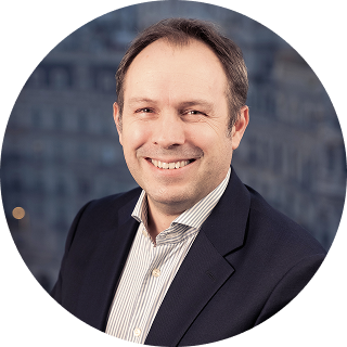 A man in a suit and striped shirt is smiling at the camera, with an office building background blurred behind him.