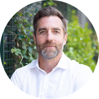 A man with short brown hair and a beard, wearing a white shirt, stands outdoors in front of green foliage and a wire fence.
