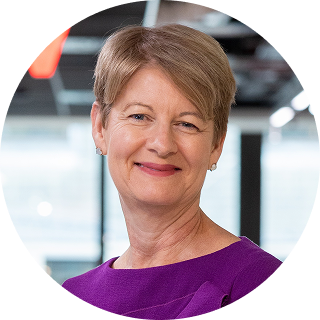 A woman with short light brown hair, wearing a purple top, smiles in an indoor office setting.