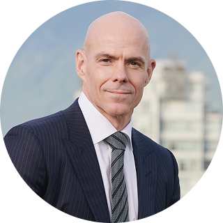 Bald man in a dark pinstripe suit and tie, standing outdoors with blurred buildings and mountains in the background.
